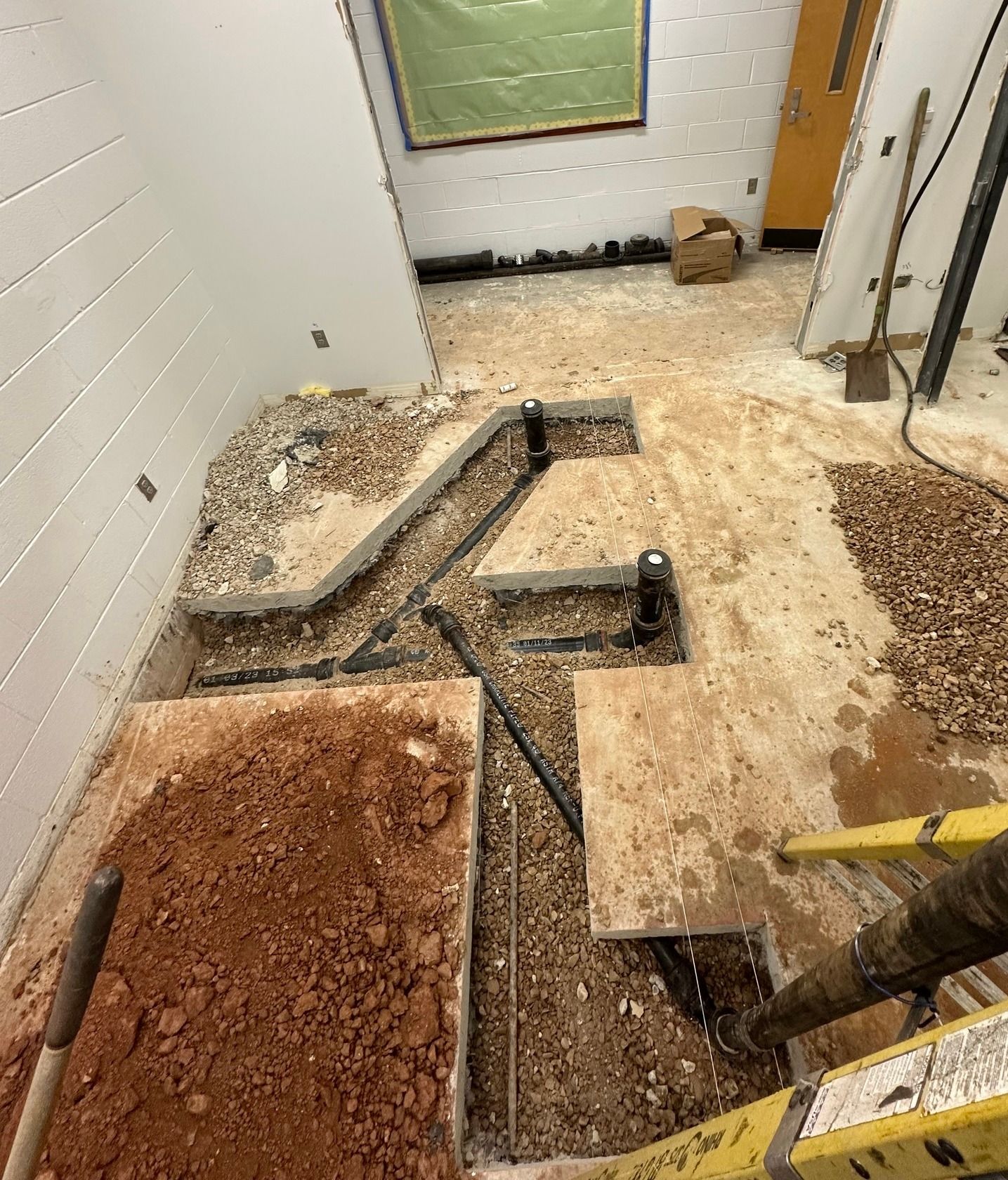 Construction site showing exposed floor plumbing trenches with black pipes, gravel, and piles of dirt.