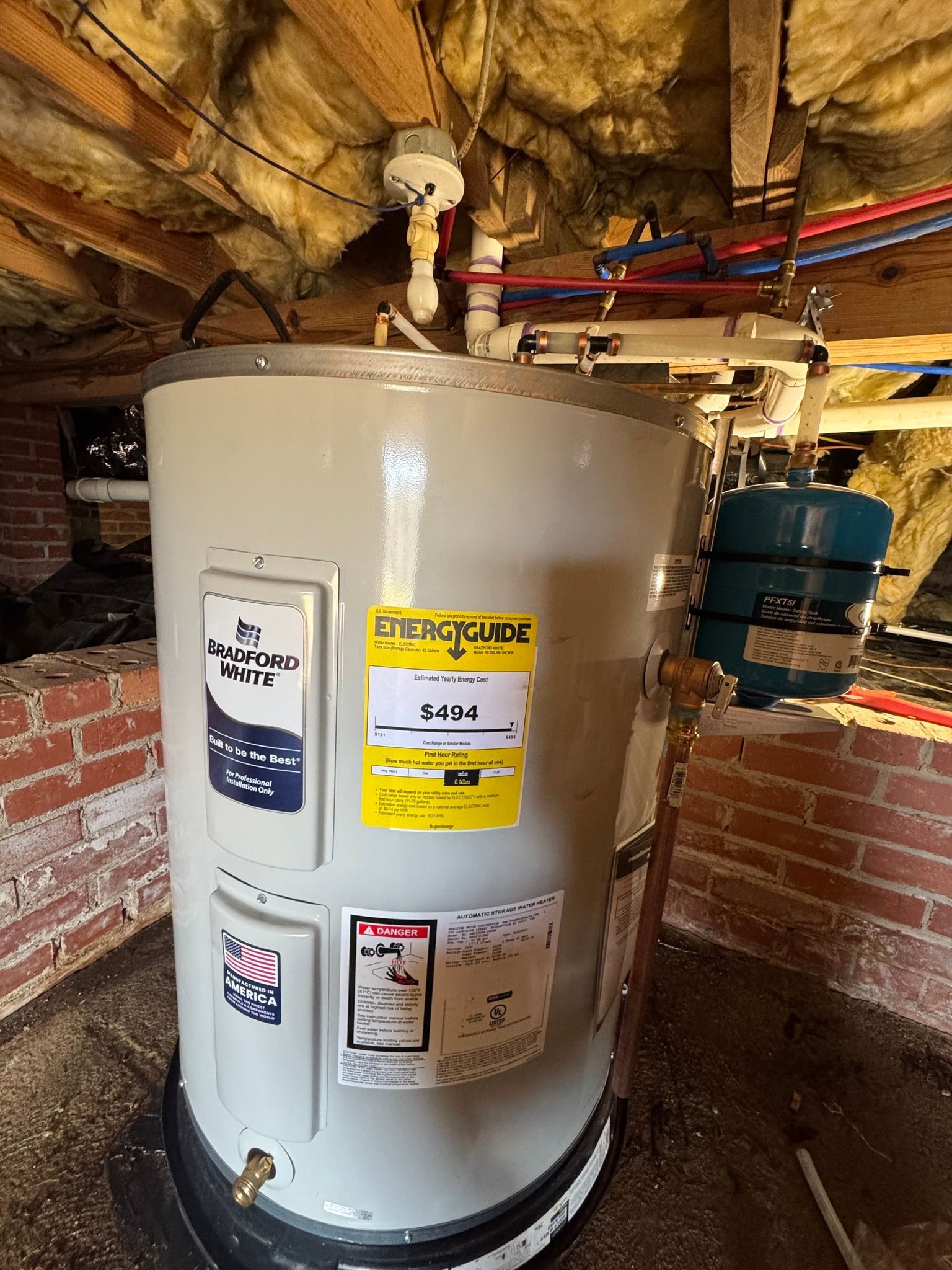 A gray electric water heater with an EnergyGuide label stands in an unfinished basement with brick walls.