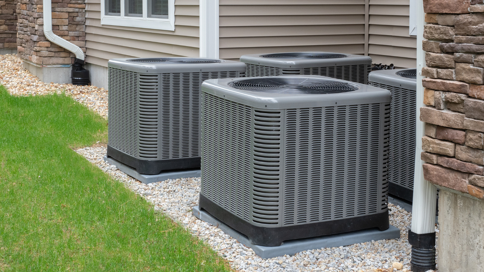 Two gray outdoor air conditioning units beside a house on a gravel bed