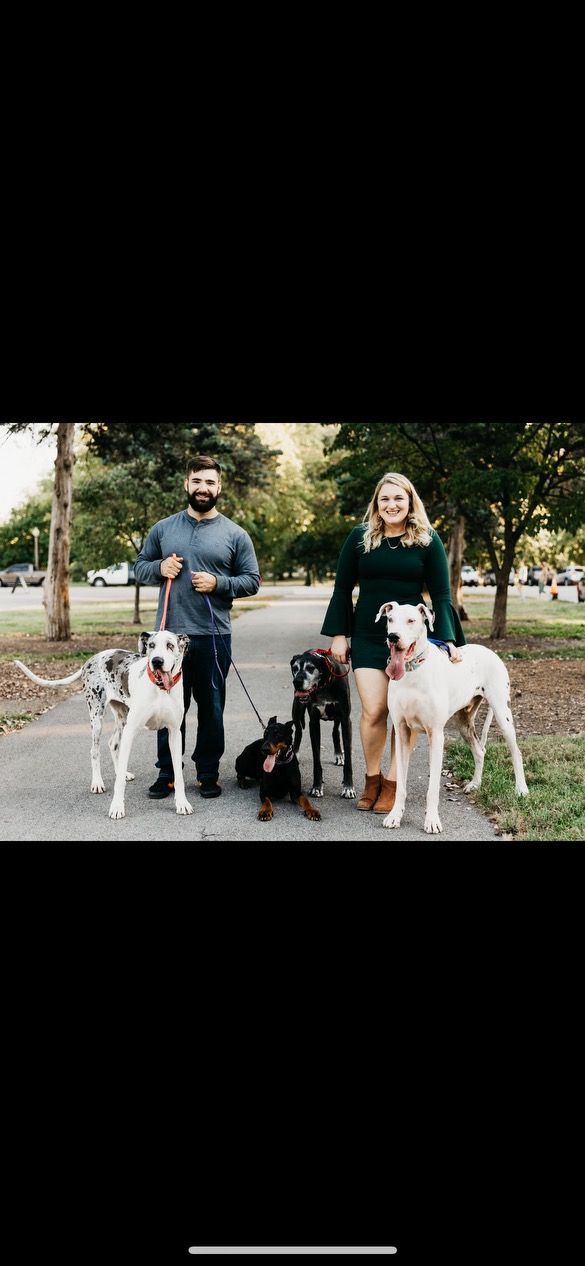 Two people stand on a path with four dogs. The dogs are on leashes. Trees are in the background.