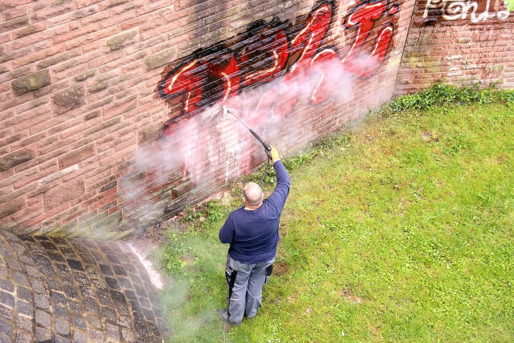 A man is cleaning graffiti on a brick wall with a high pressure washer.