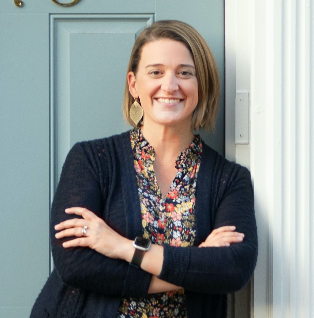 Woman with arms crossed, smiling, wearing floral shirt, cardigan, and earrings, leaning against a blue door.