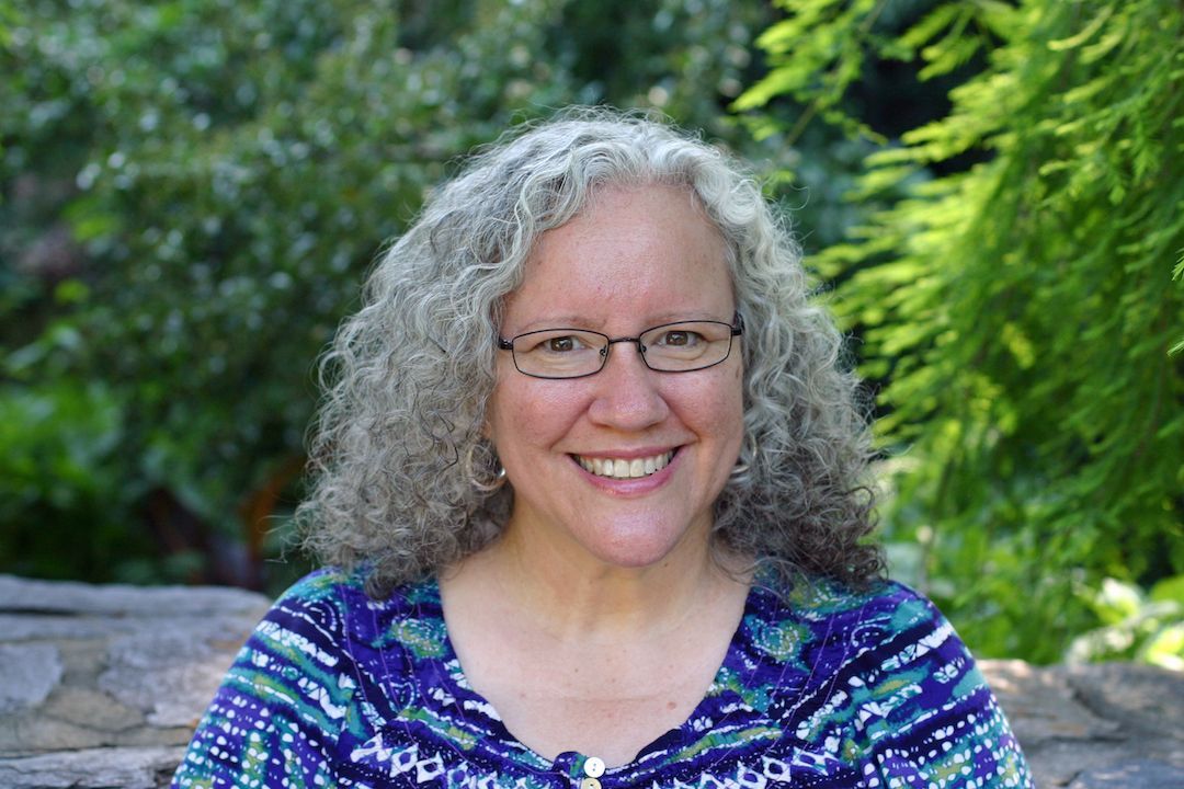 Woman with gray curly hair and glasses smiles outdoors.