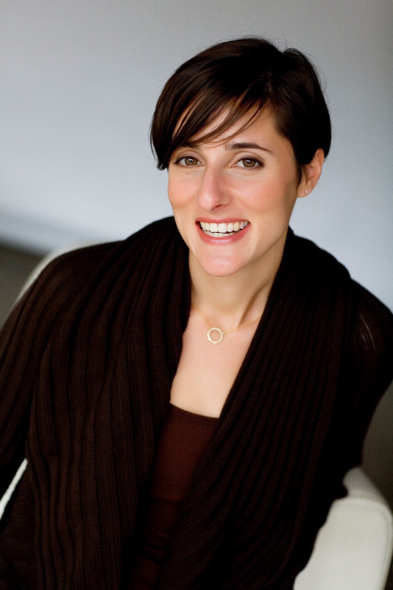 Woman with short dark hair, smiling, wearing a brown sweater and a gold necklace.