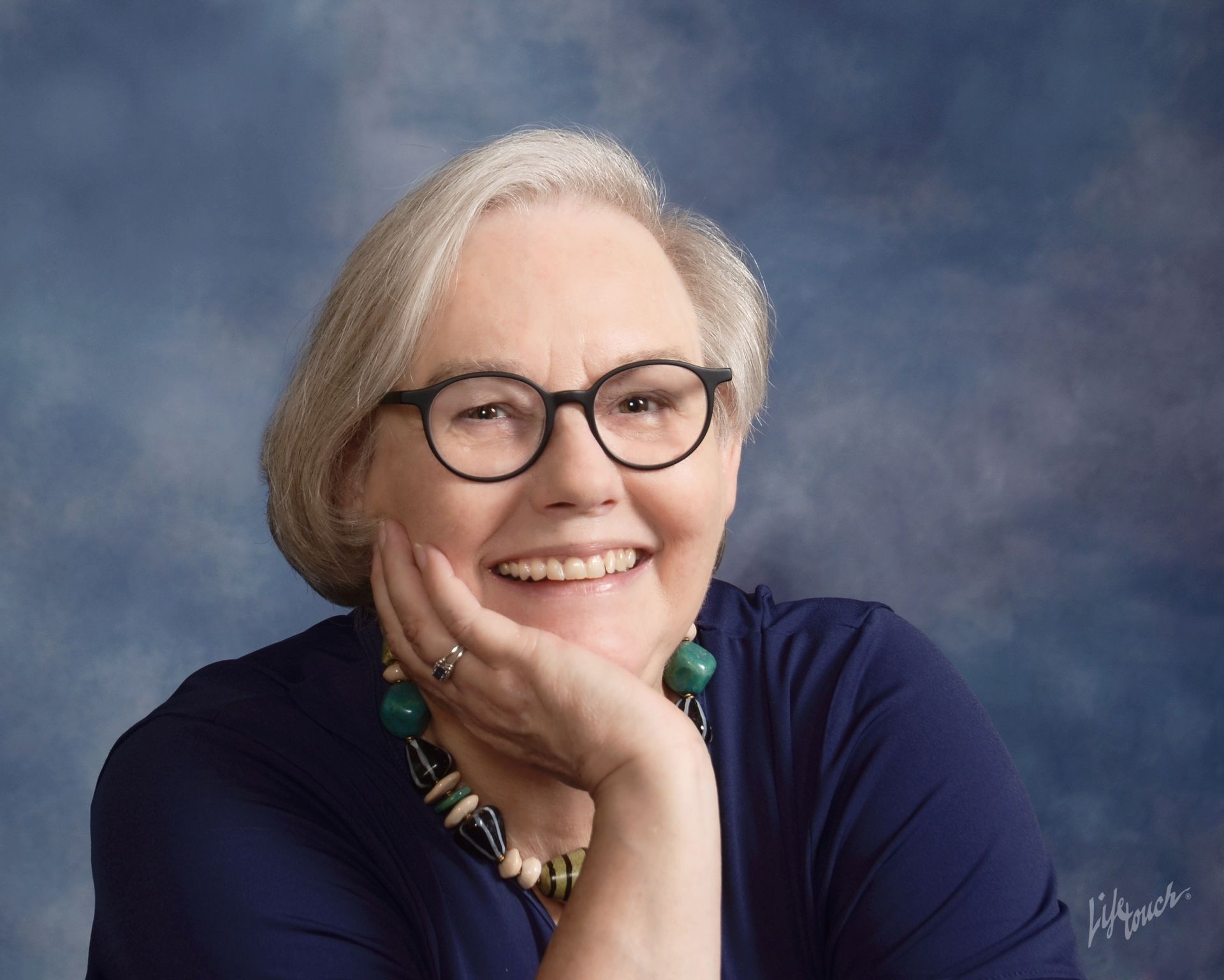 Woman with glasses smiles, resting chin on hand; blue top, beaded necklace; blue and cloudy backdrop.