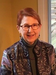 Woman with short red hair and glasses smiles. Wearing a patterned jacket and olive turtleneck. Indoor setting.
