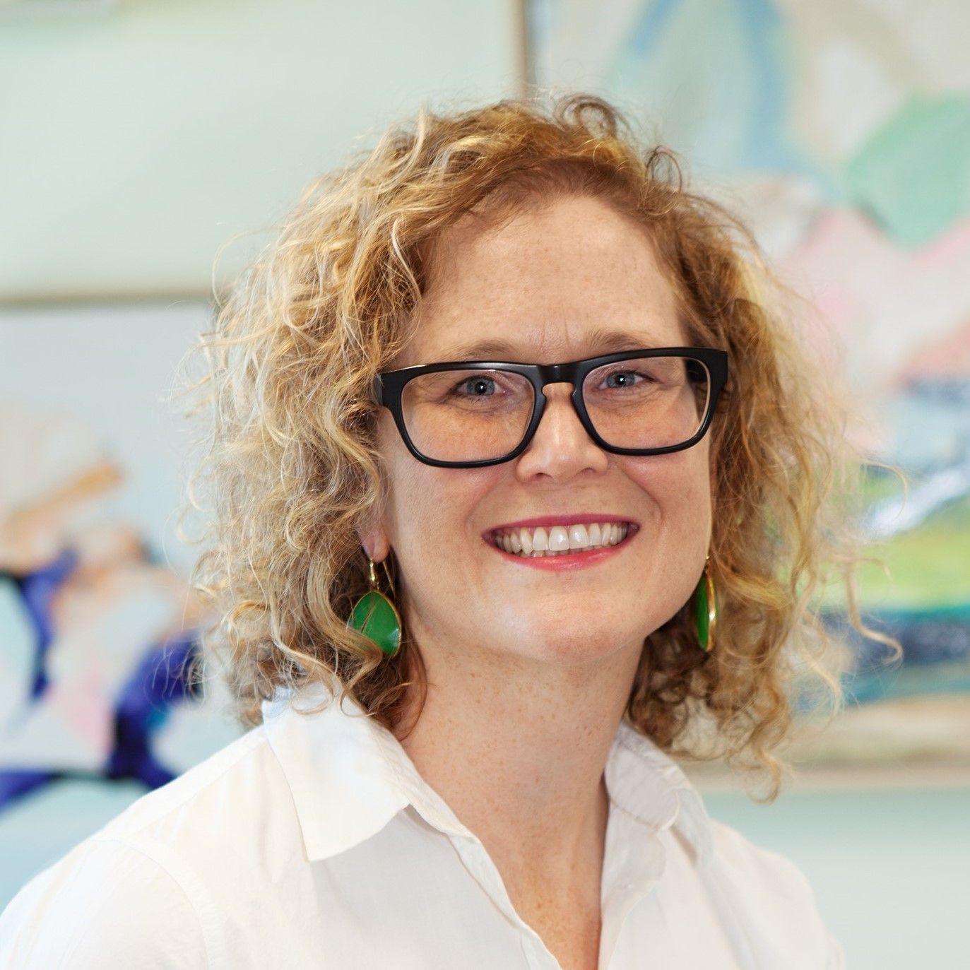 Woman with curly blonde hair and glasses smiles. Wearing green earrings and a white shirt.