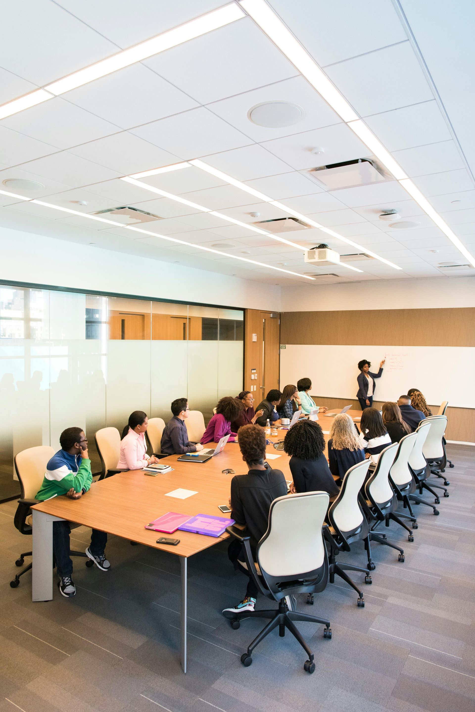 People in a meeting at a long table; a person stands at a whiteboard, presenting. Brightly lit room.