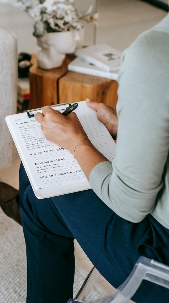 Person writing on a clipboard, sitting, legs crossed, in a room with a side table, with a potted plant on top.