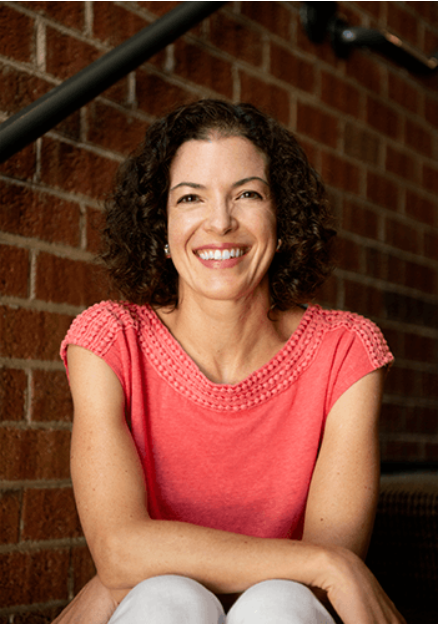 Woman with curly hair smiles, seated on steps against brick wall; wears coral top, arms crossed.