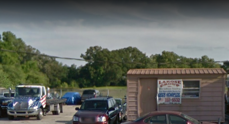 Car lot with shed, cars, tow truck; green trees and bridge in background.