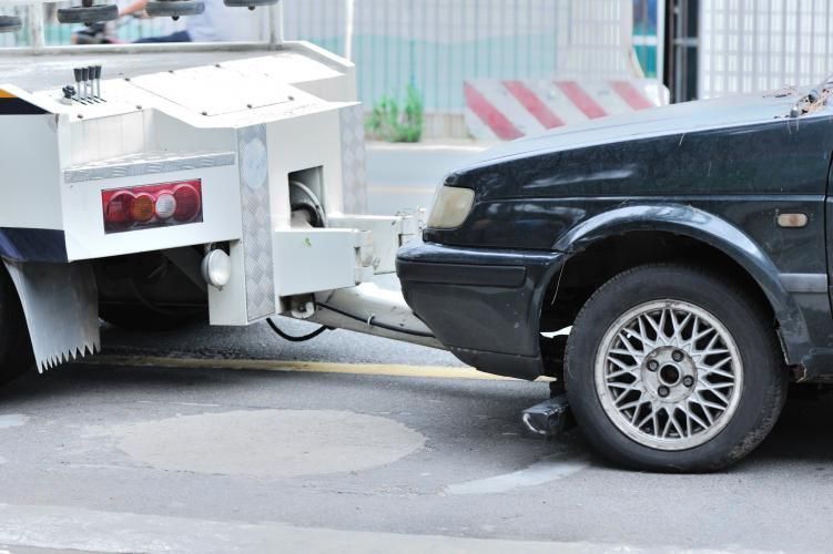 Tow truck towing a black car on a street, the front of the car is damaged.