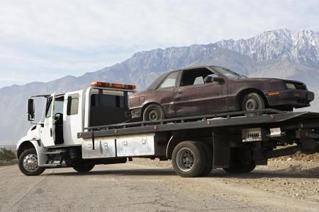 Tow truck carrying a damaged, brown car on a roadside with mountains in the background.