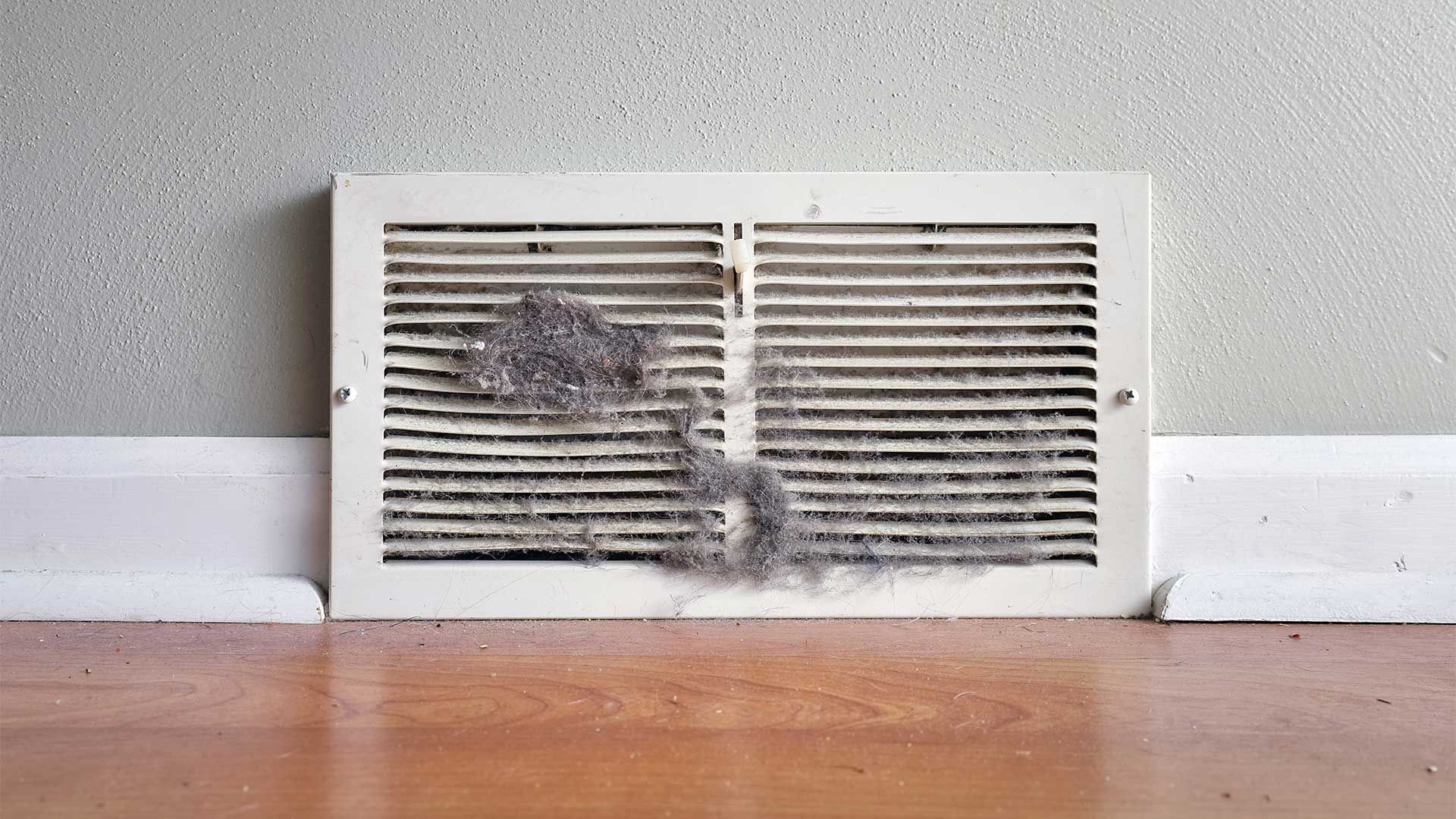 A dirty air vent is sitting on a wooden floor next to a wall.