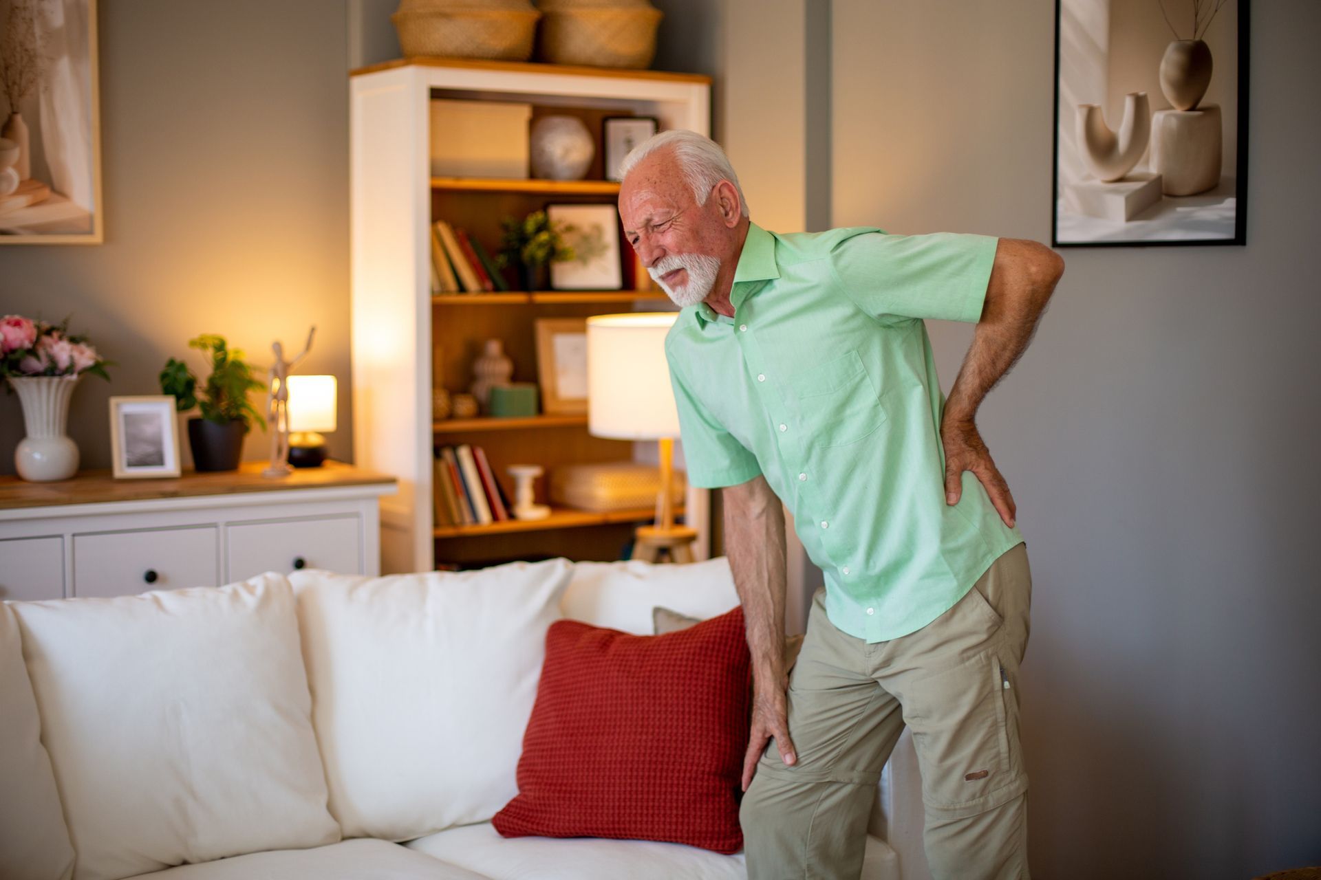 Elderly man holds lower back in pain beside white living room couch.