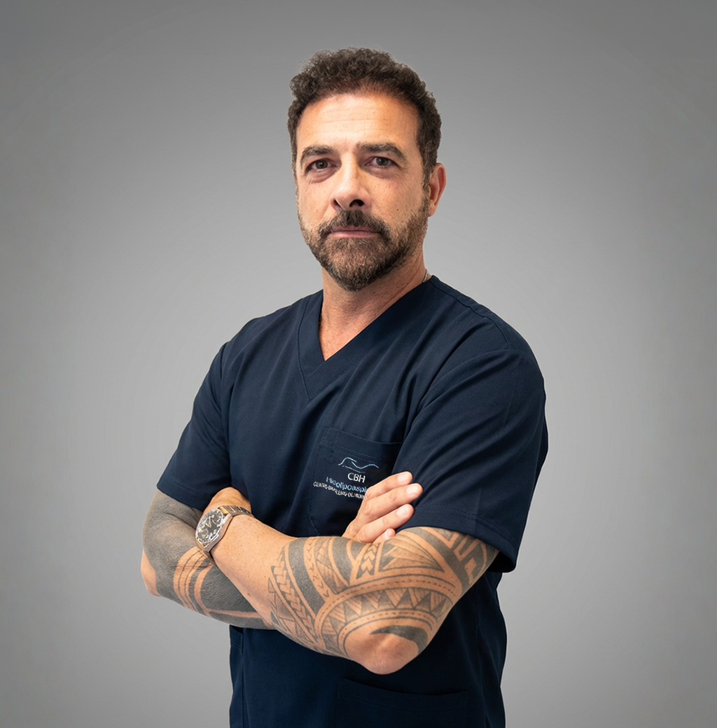 Um homem com barba e tatuagens está de pé com os braços cruzados.
