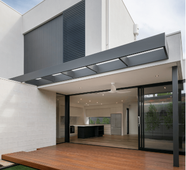 Exterior de casa moderna com portas de correr abertas que levam à cozinha. Deck de madeira escura e toldo cinza.