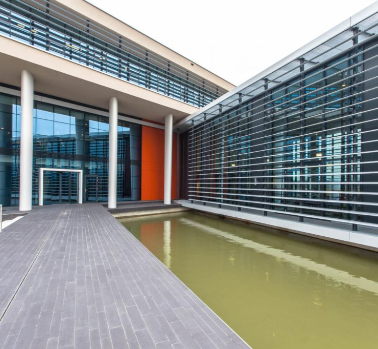 Exterior de edifício moderno com paredes de vidro, uma longa passarela sobre a água e uma parede com detalhes em laranja.