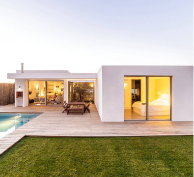 Casa branca moderna com piscina, deck e gramado. Quarto e sala de estar visíveis. Mesa e cadeiras de madeira.