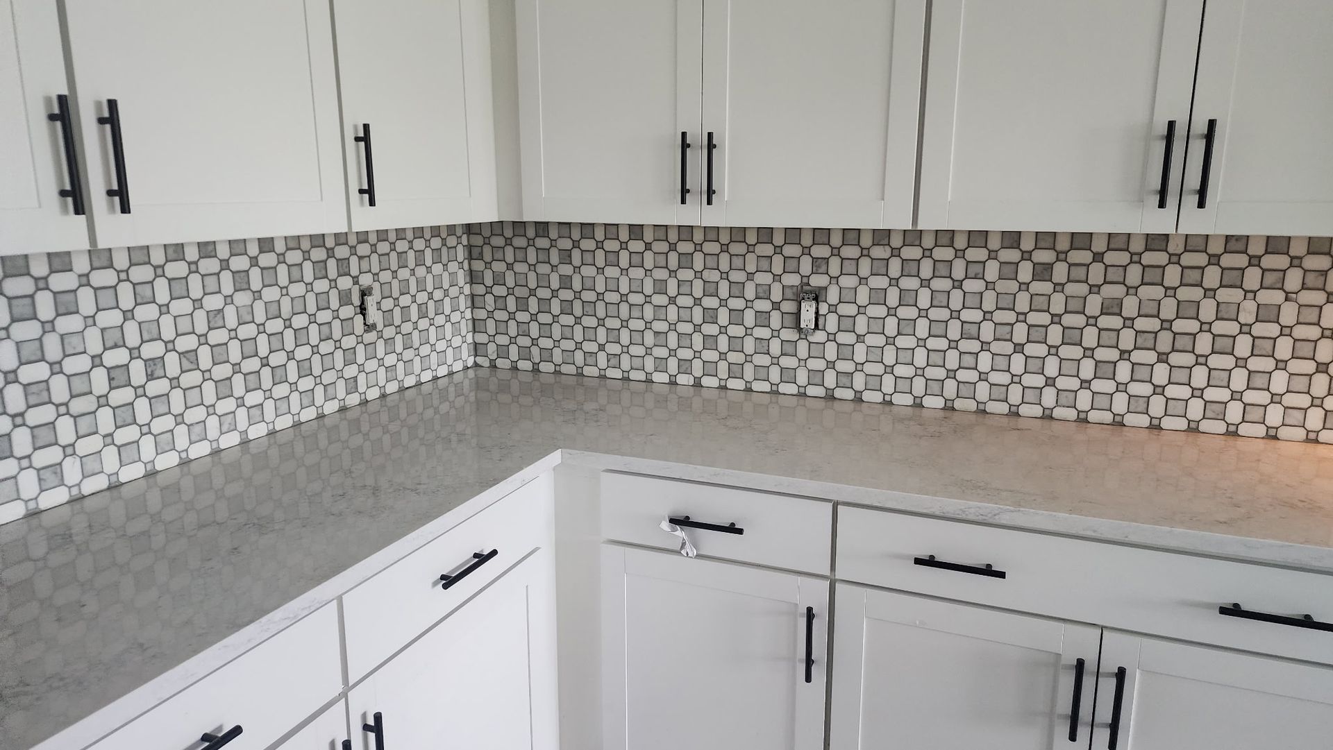 White kitchen cabinets with black handles and a gray countertop, mosaic backsplash.