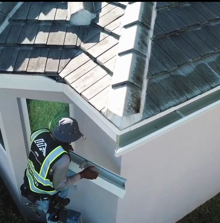 Roofer installing gutter on a light-colored house with dark roof tiles. He is wearing a safety vest.
