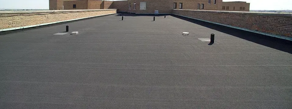 A flat, black rooftop with multiple vents; a building is visible in the background.