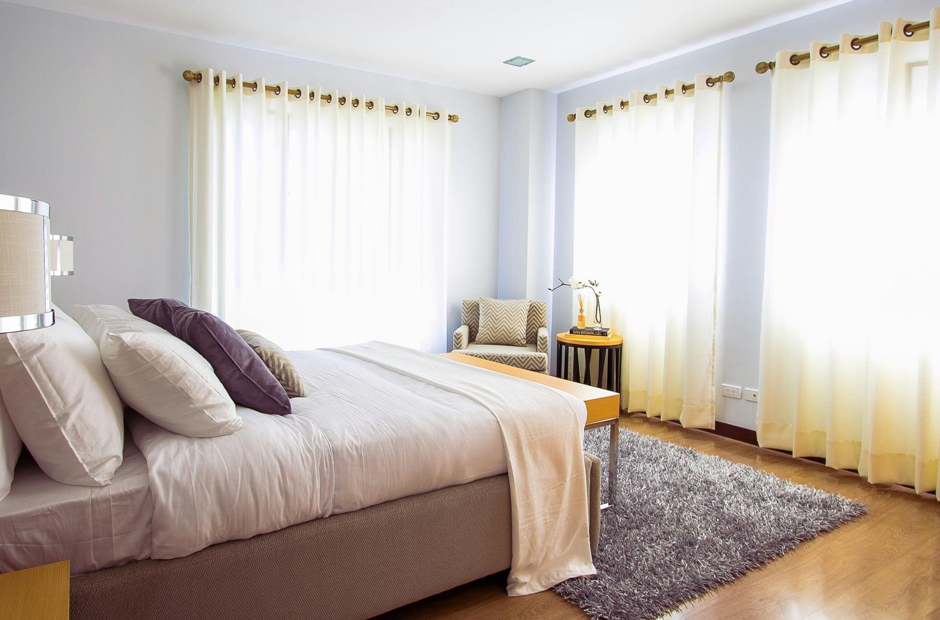 Bedroom with a bed, pillows, chair, and windows with curtains; light blue walls, wooden floor, and a fluffy gray rug.