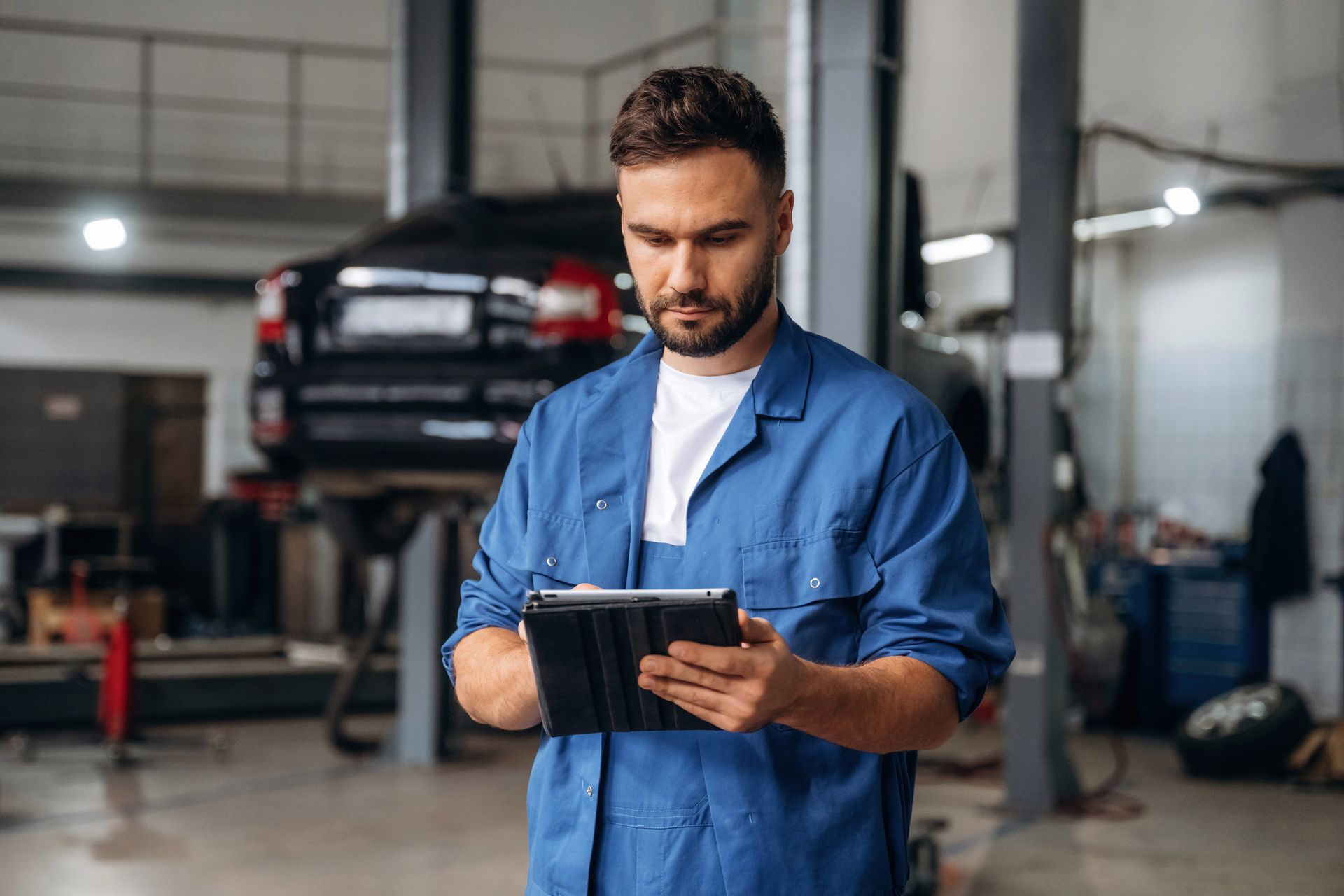 A mechanic in a blue uniform stands in an auto repair shop, looking down at a tablet while holding a stylus. A mechanic in a blue uniform stands in an auto repair shop, looking down at a tablet while holding a stylus.