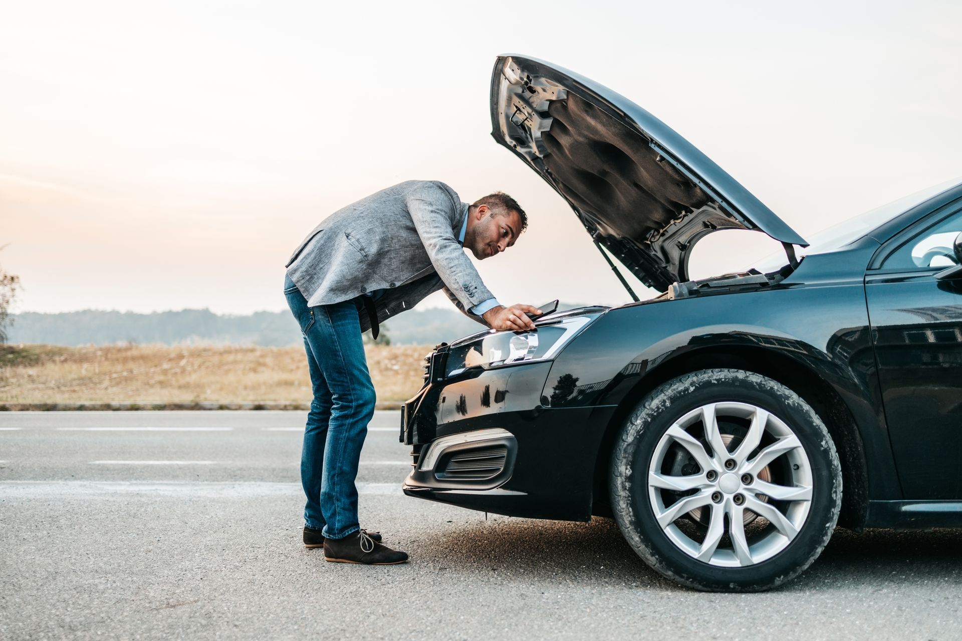 A person in business casual attire inspects the engine of a black car parked on the side of a road with the hood up. A person in business casual attire inspects the engine of a black car parked on the side of a road with the hood up.