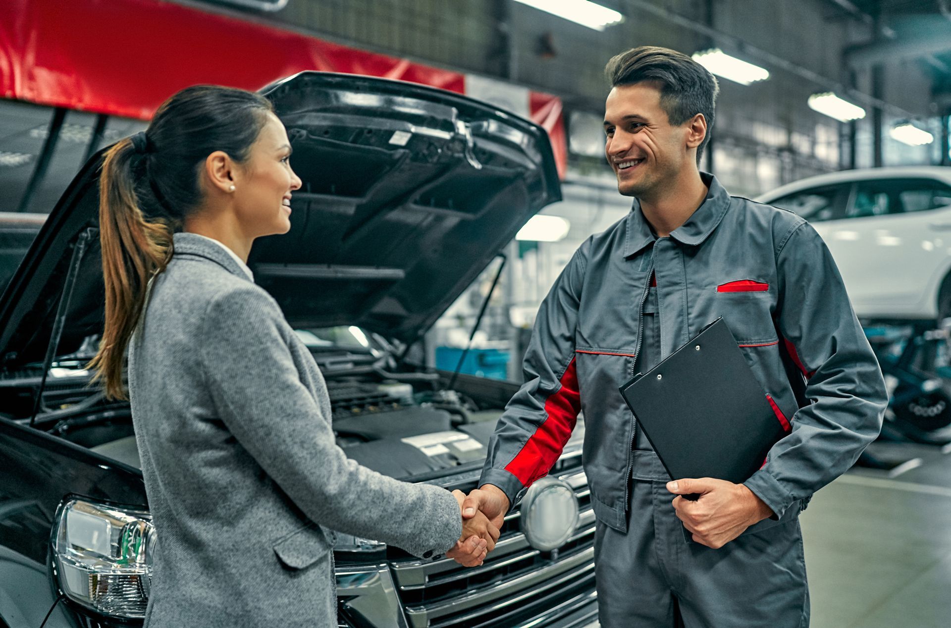 A service professional in a grey uniform shakes hands with a customer in front of a car with an open hood in a garage. A service professional in a grey uniform shakes hands with a customer in front of a car with an open hood in a garage.