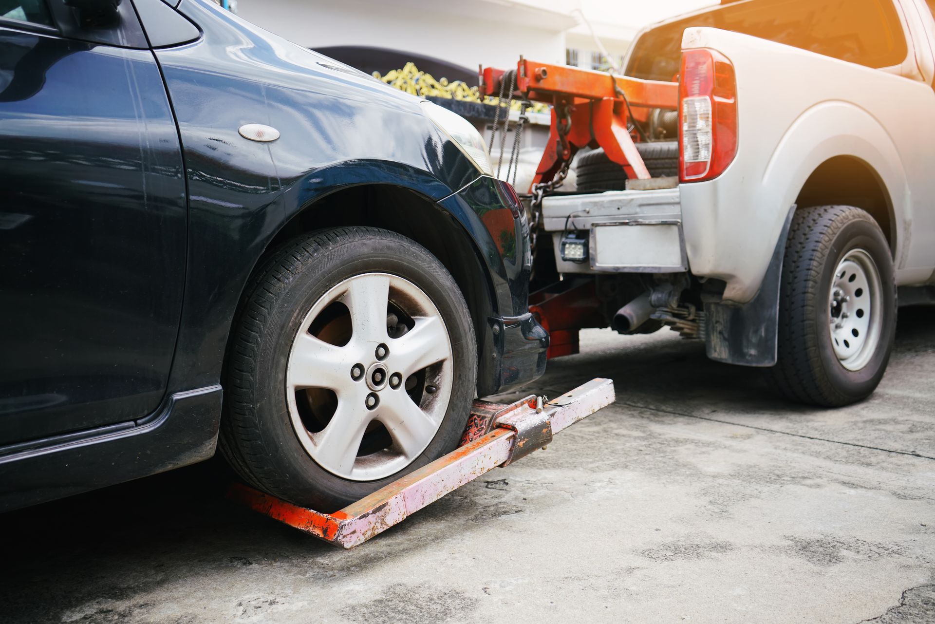 A silver tow truck with an orange wheel lift is hooked to the front tire of a black car on a paved surface. A silver tow truck with an orange wheel lift is hooked to the front tire of a black car on a paved surface.