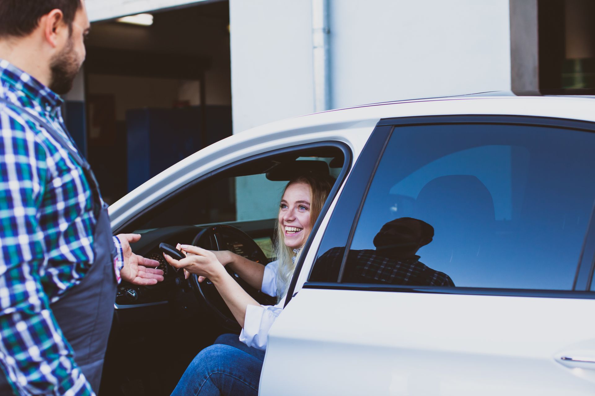 A smiling person sitting in a white car hands a set of keys to a mechanic wearing a plaid shirt and overalls. A smiling person sitting in a white car hands a set of keys to a mechanic wearing a plaid shirt and overalls.