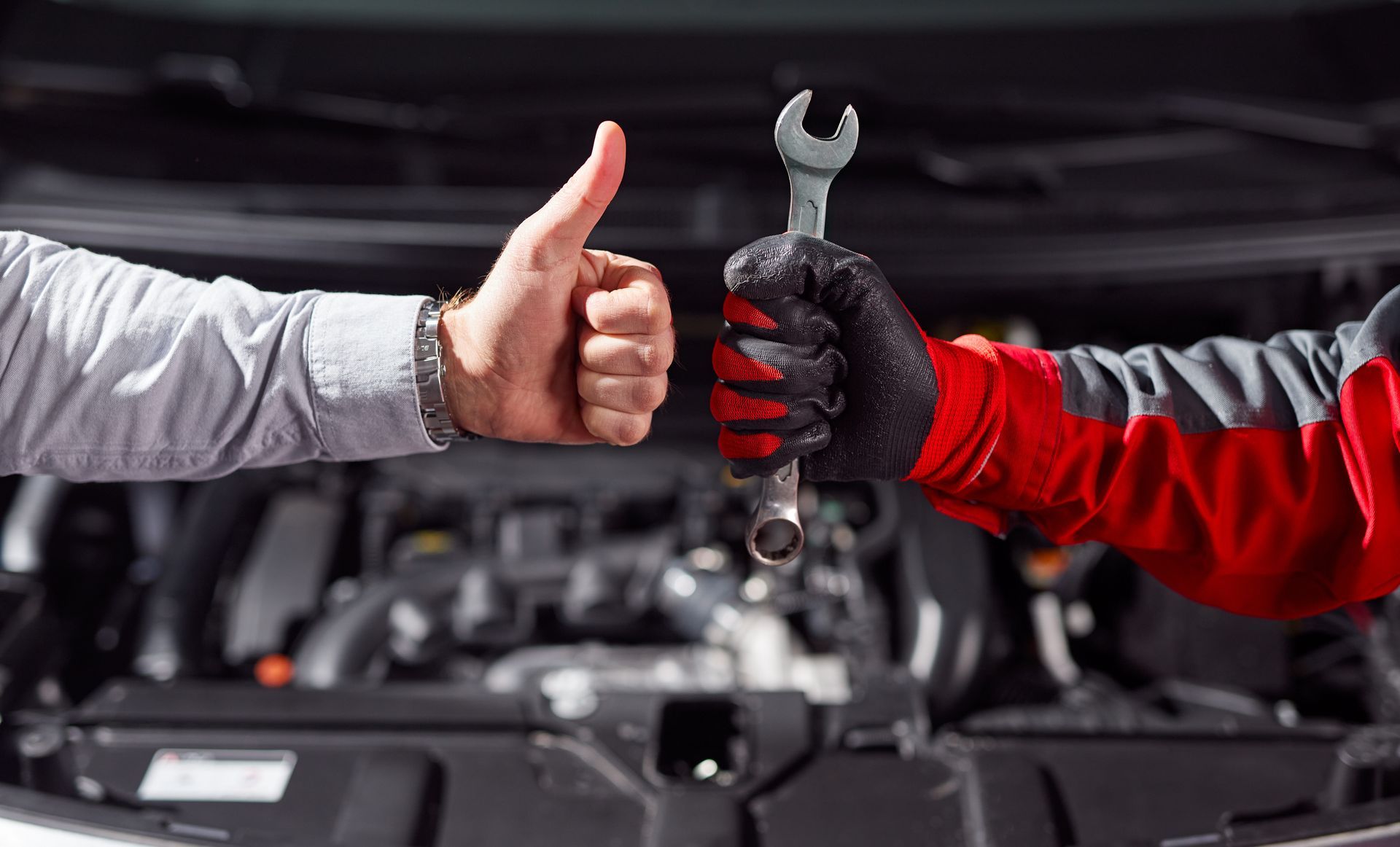 A customer gives a thumbs up to a mechanic holding a wrench in front of an open car engine. A customer gives a thumbs up to a mechanic holding a wrench in front of an open car engine.