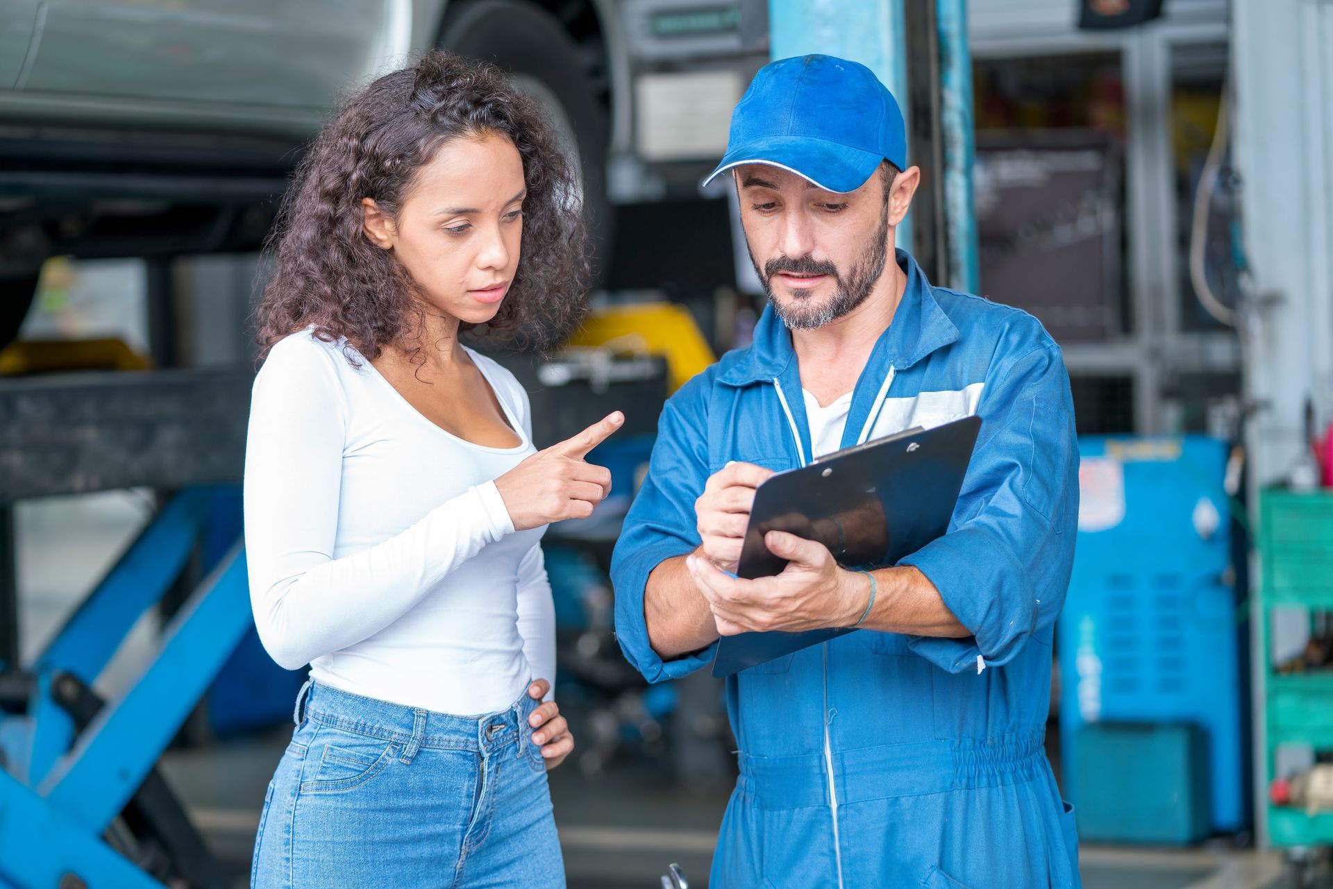A mechanic in blue coveralls and a cap discusses a clipboard with a client inside a vehicle repair shop. A mechanic in blue coveralls and a cap discusses a clipboard with a client inside a vehicle repair shop.