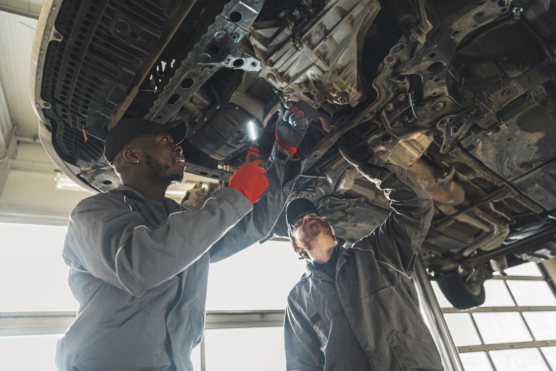 Two mechanics in grey uniforms work under a car on a lift, using a flashlight to inspect the undercarriage in a garage.