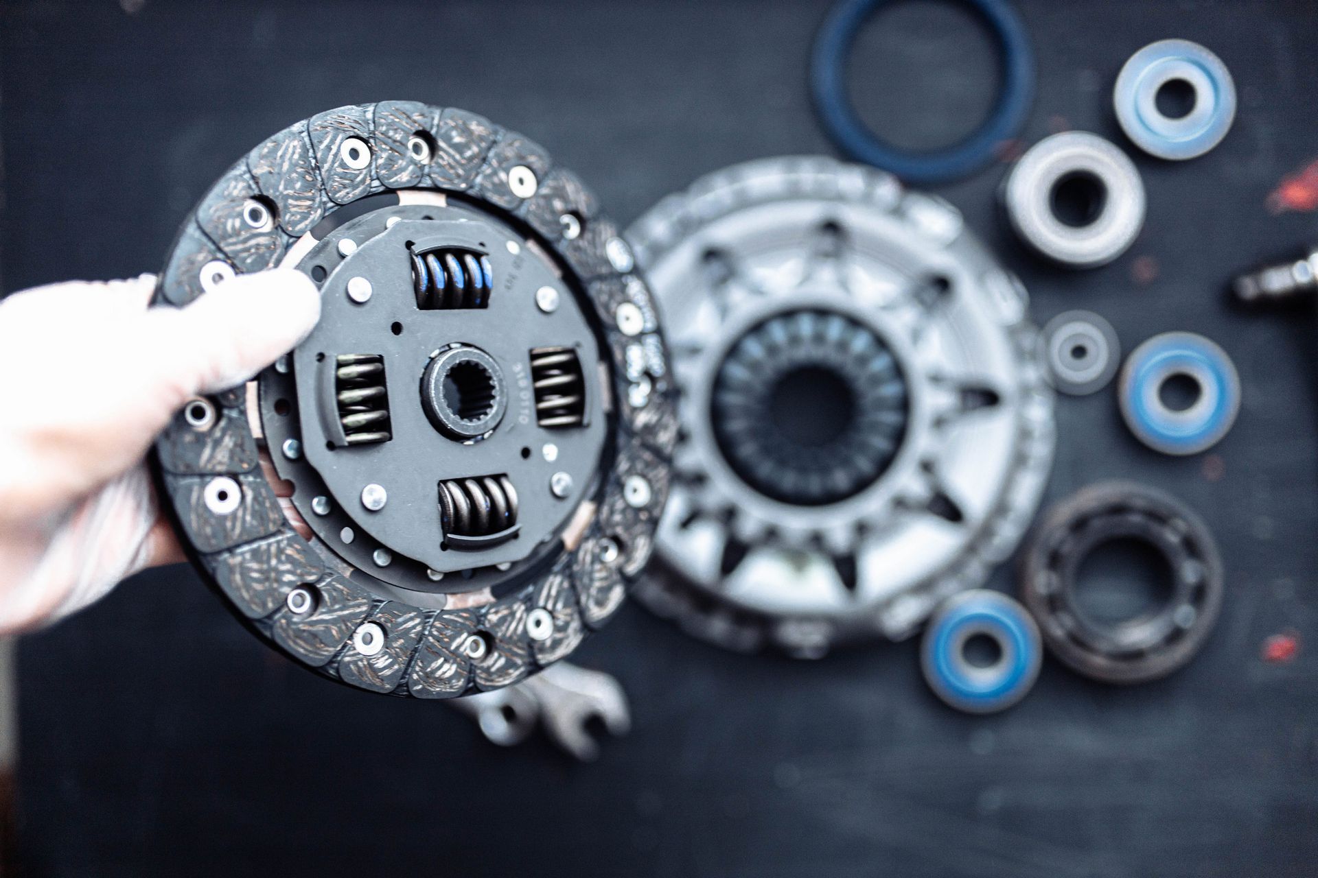 A gloved hand holds a car clutch disc above a work surface containing a clutch pressure plate and several bearings.
