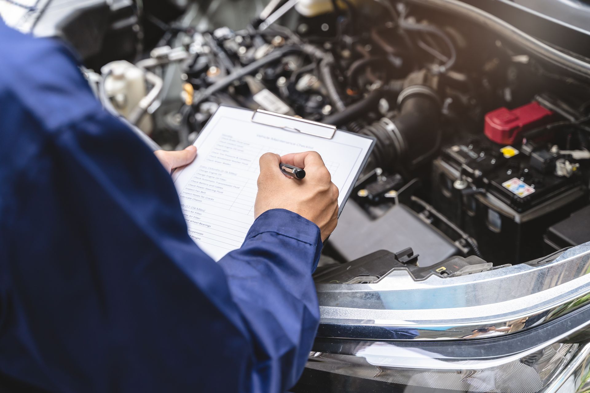 A mechanic in a blue uniform writes on a clipboard while inspecting an open car engine. A mechanic in a blue uniform writes on a clipboard while inspecting an open car engine.