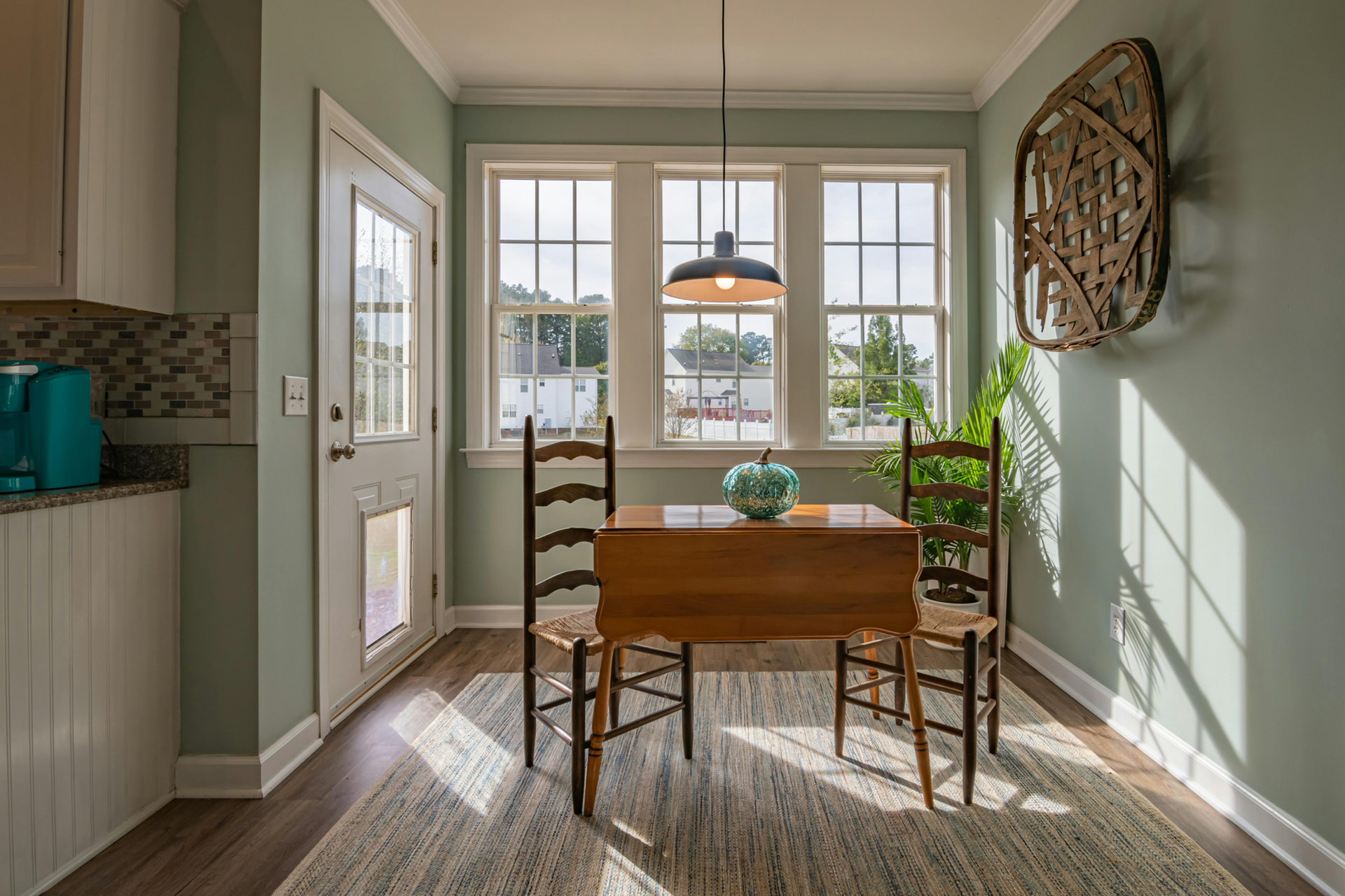 Breakfast nook with table, chairs, windows, and a woven wall decoration. Pale green walls and sunlight.