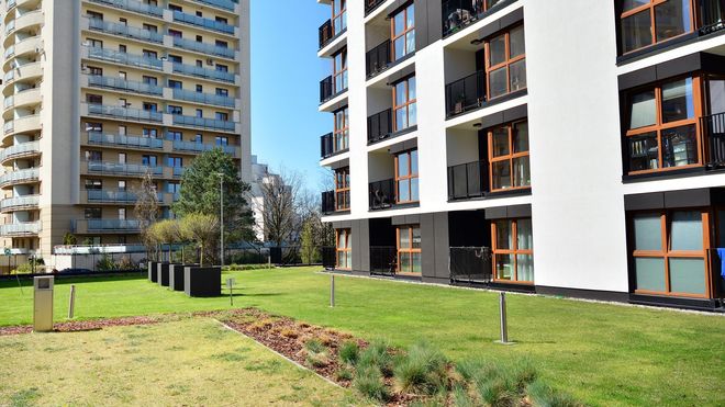 Apartment buildings with balconies and windows, next to a grassy lawn under a blue sky.