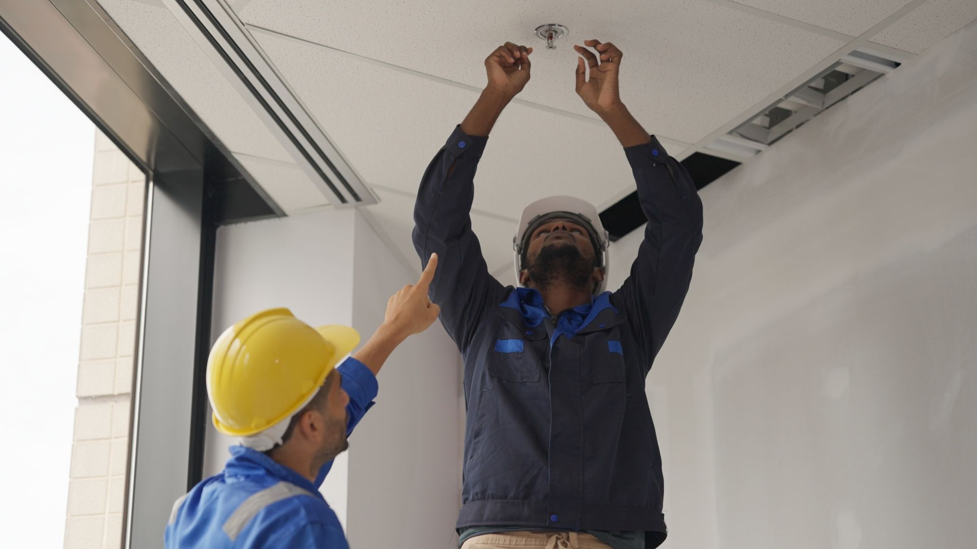 Two workers in hard hats inspecting or adjusting a ceiling fixture in a white room
