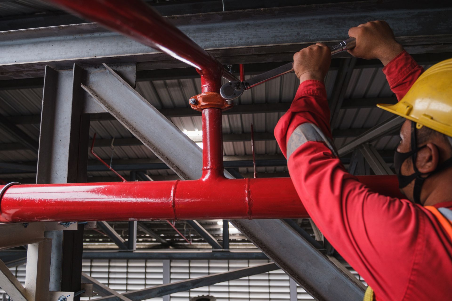 Worker in red jacket and yellow hard hat installing red pipes in an industrial ceiling.