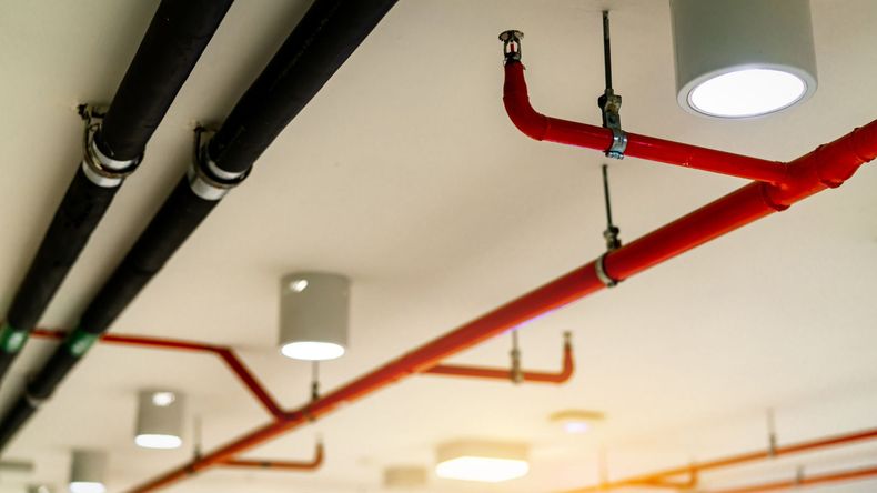 Ceiling view of red and black exposed pipes with white light fixtures in an indoor space