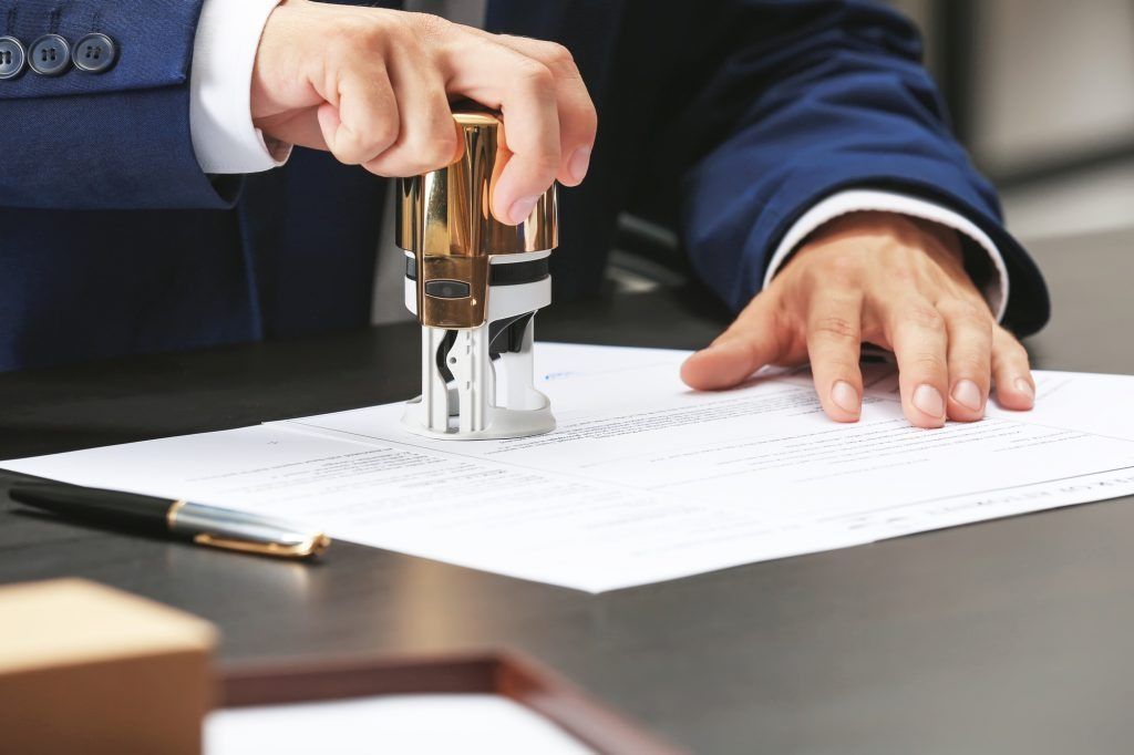 Hand stamping a document on a desk beside a pen and laptop.