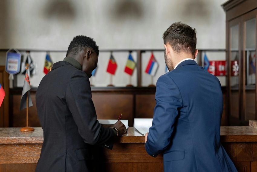 Two men in suits signing a document at a table, with small national flags displayed behind them.