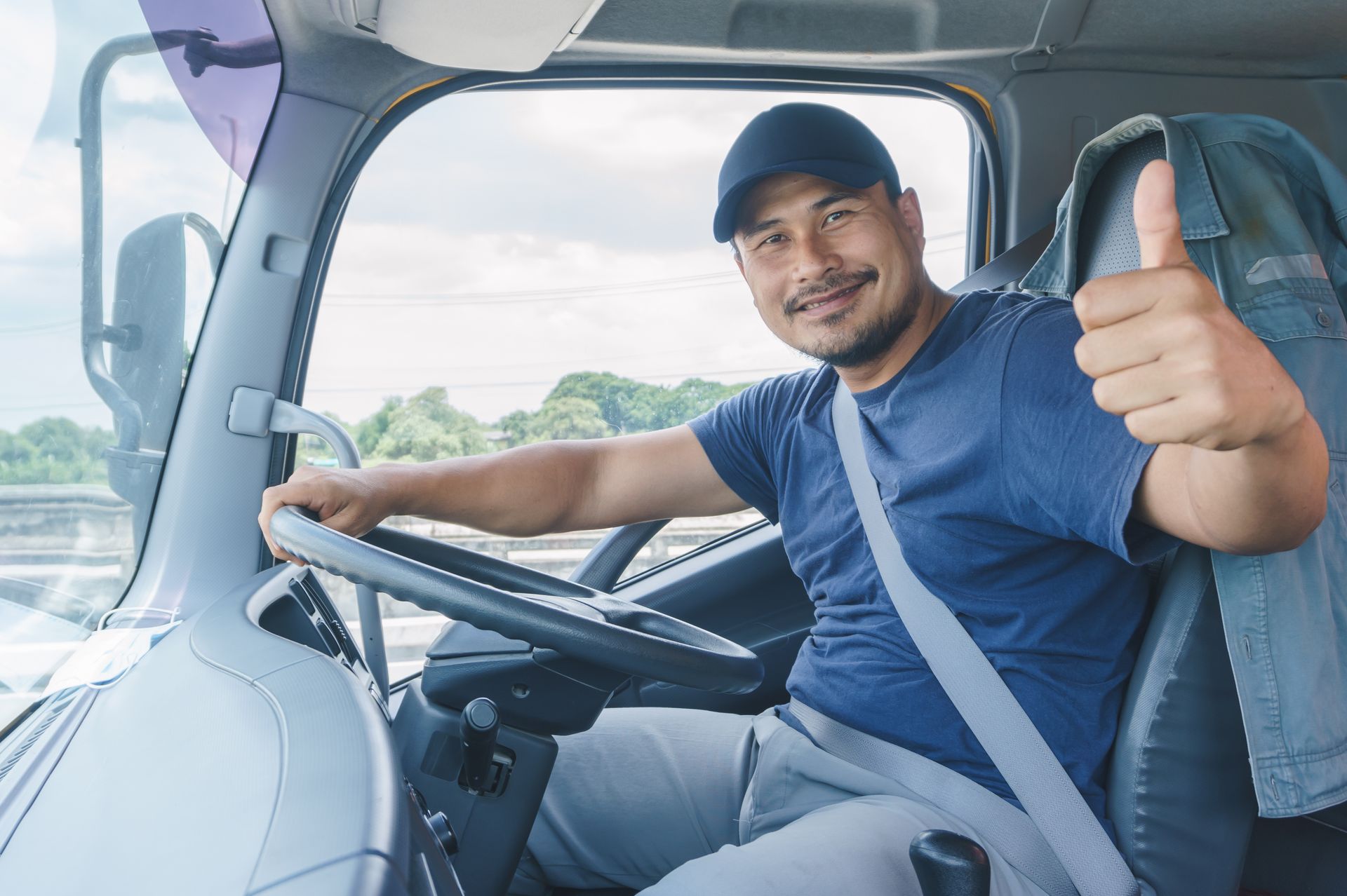 Smiling truck driver giving a thumbs-up inside a diesel truck, wearing a seatbelt.
