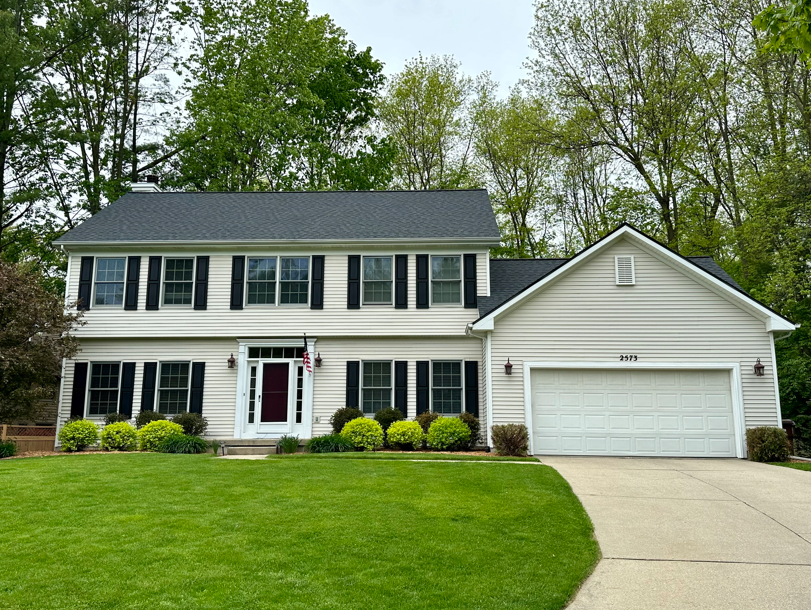 Two-story beige house with black shutters, black roof, and attached garage; green lawn and trees.