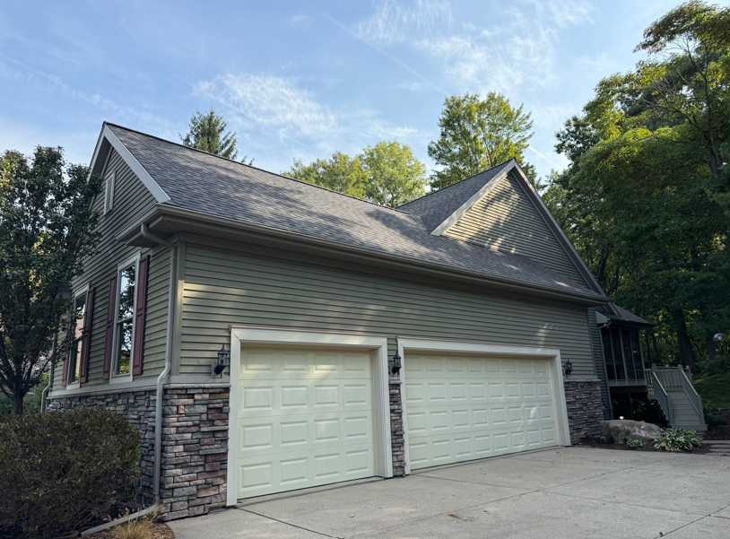 Two-car garage with light tan doors and green siding. Stone accents around garage and base. Roof is gray.
