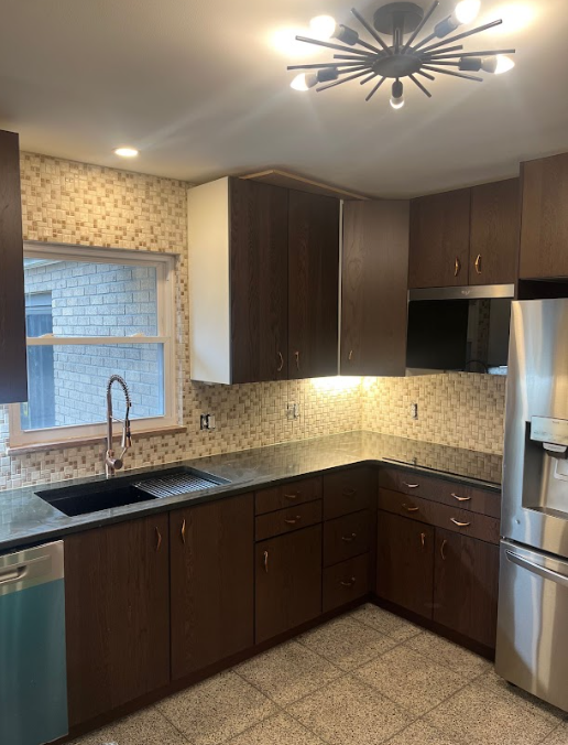 Dark brown kitchen cabinets, stainless steel refrigerator, black sink, patterned backsplash and light granite countertops.