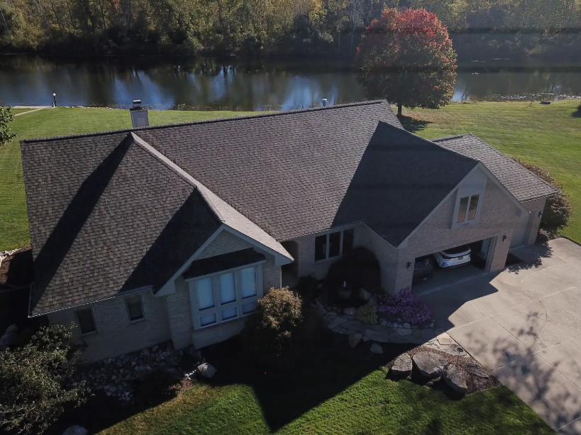House with brown shingle roof, facing lake. Green lawn, trees in background, car in garage.