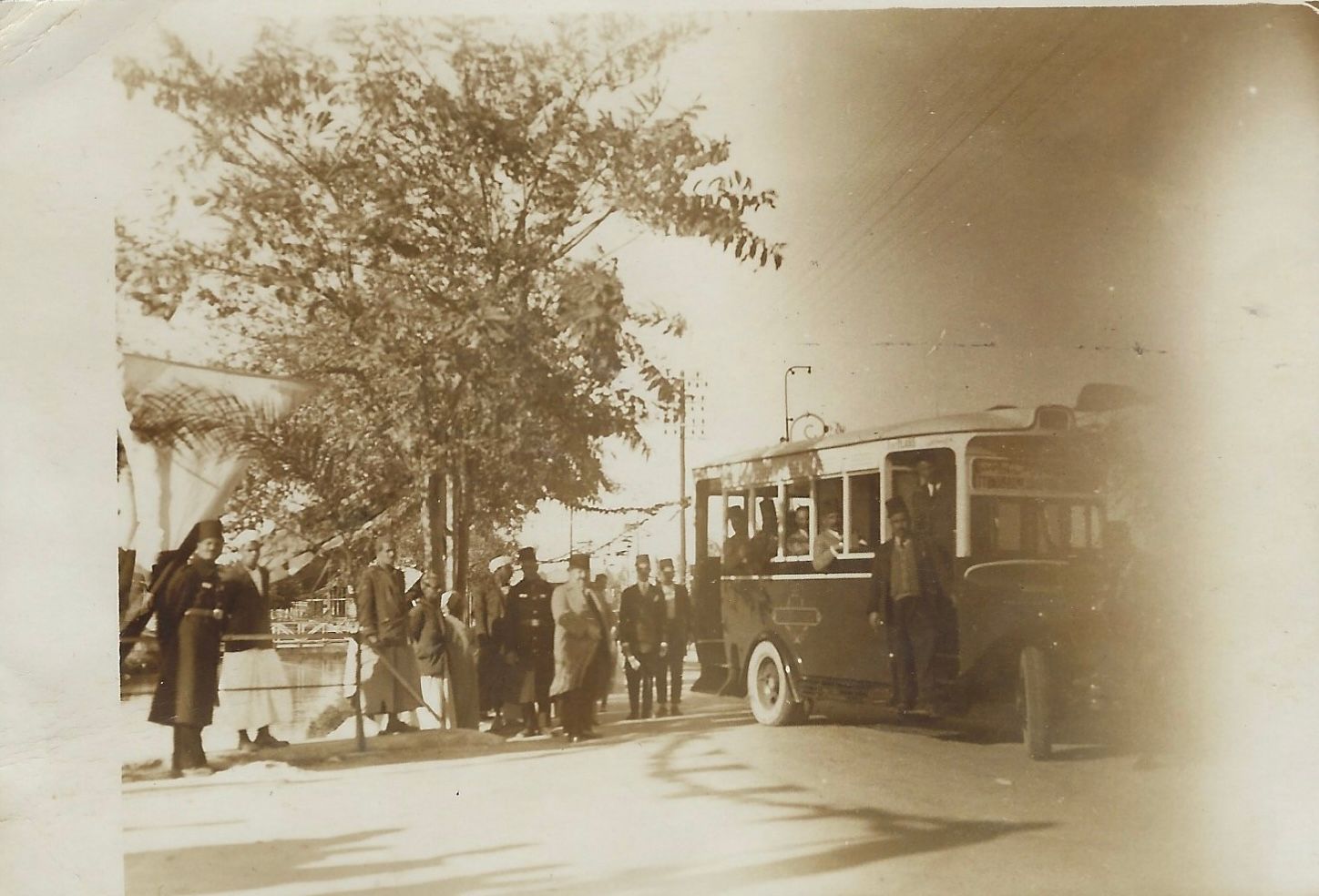 People boarding a trolley bus, near a tree and building. Sepia-toned, likely a historical photo.